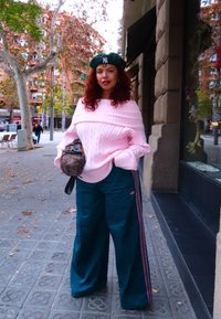 Femme aux cheveux roux bouclés portant un béret vert, un pull rose épaules dénudées, un pantalon large émeraude, tenant un sac à main à motif sur un trottoir de ville en automne.