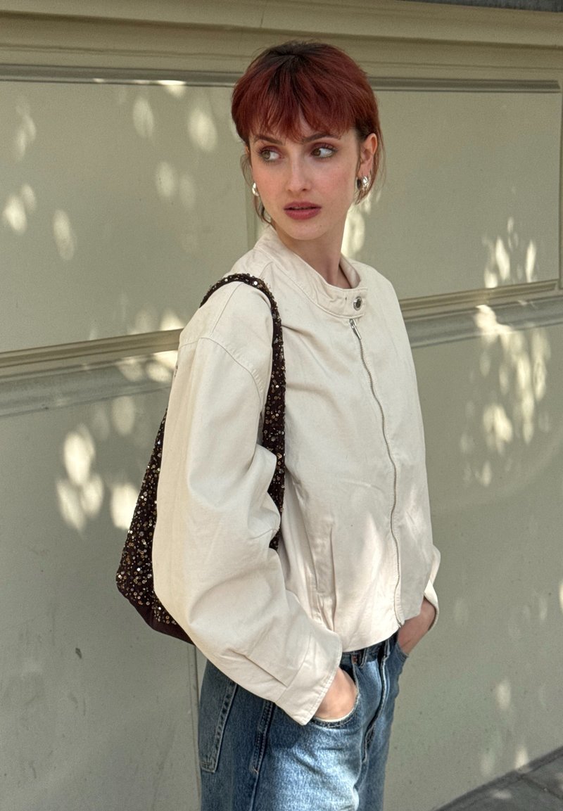 Personne aux cheveux roux portant une veste beige clair et un jean bleu, debout contre un mur clair avec les mains dans les poches, regardant sur le côté, portant un sac à motif.