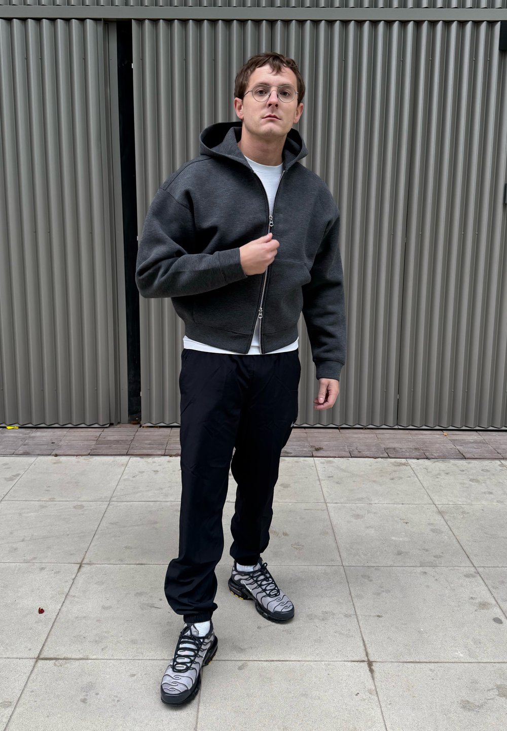 Camisola com capuz cinzento escuro com fecho de correr, combinada com uma camisa branca, calças pretas e sapatos cinzentos com detalhes em preto e amarelo.