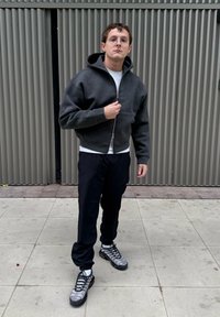 Camisola com capuz cinzento escuro com fecho de correr, combinada com uma camisa branca, calças pretas e sapatos cinzentos com detalhes em preto e amarelo.