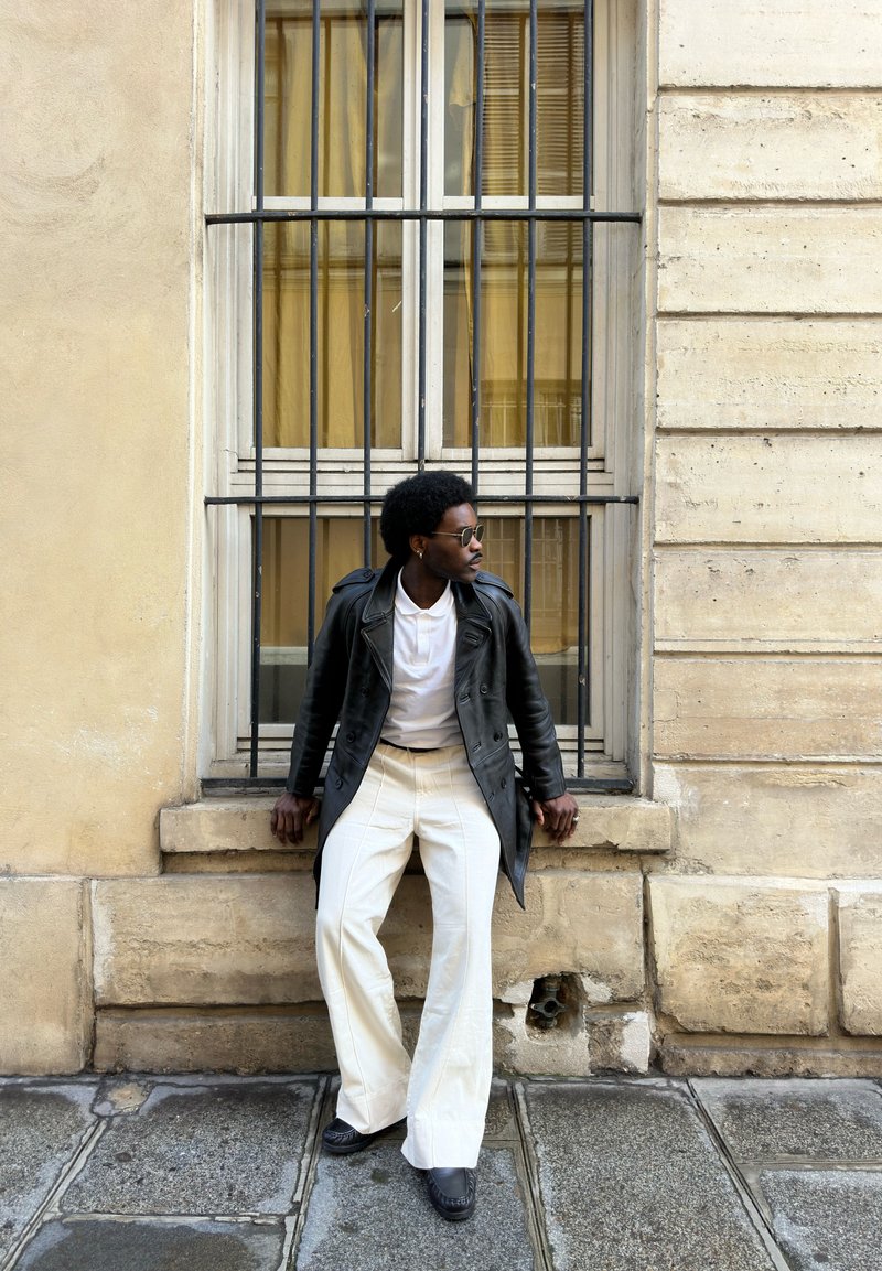 Homme portant des lunettes de soleil, un manteau en cuir noir et un pantalon blanc, appuyé contre une fenêtre grillagée sur une façade en pierre beige.