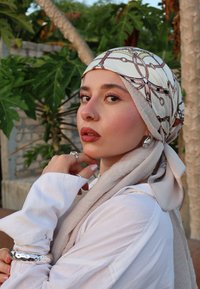 Κεφαλομάντηλο με σχέδιο σε ανοιχτόχρωμο φόντο σε κρέμα, με λεπτομέρειες σε καφέ και χρυσό. Λευκό πουκάμισο με μακρύ μανίκι και οραστά ασημένια κοσμήματα.