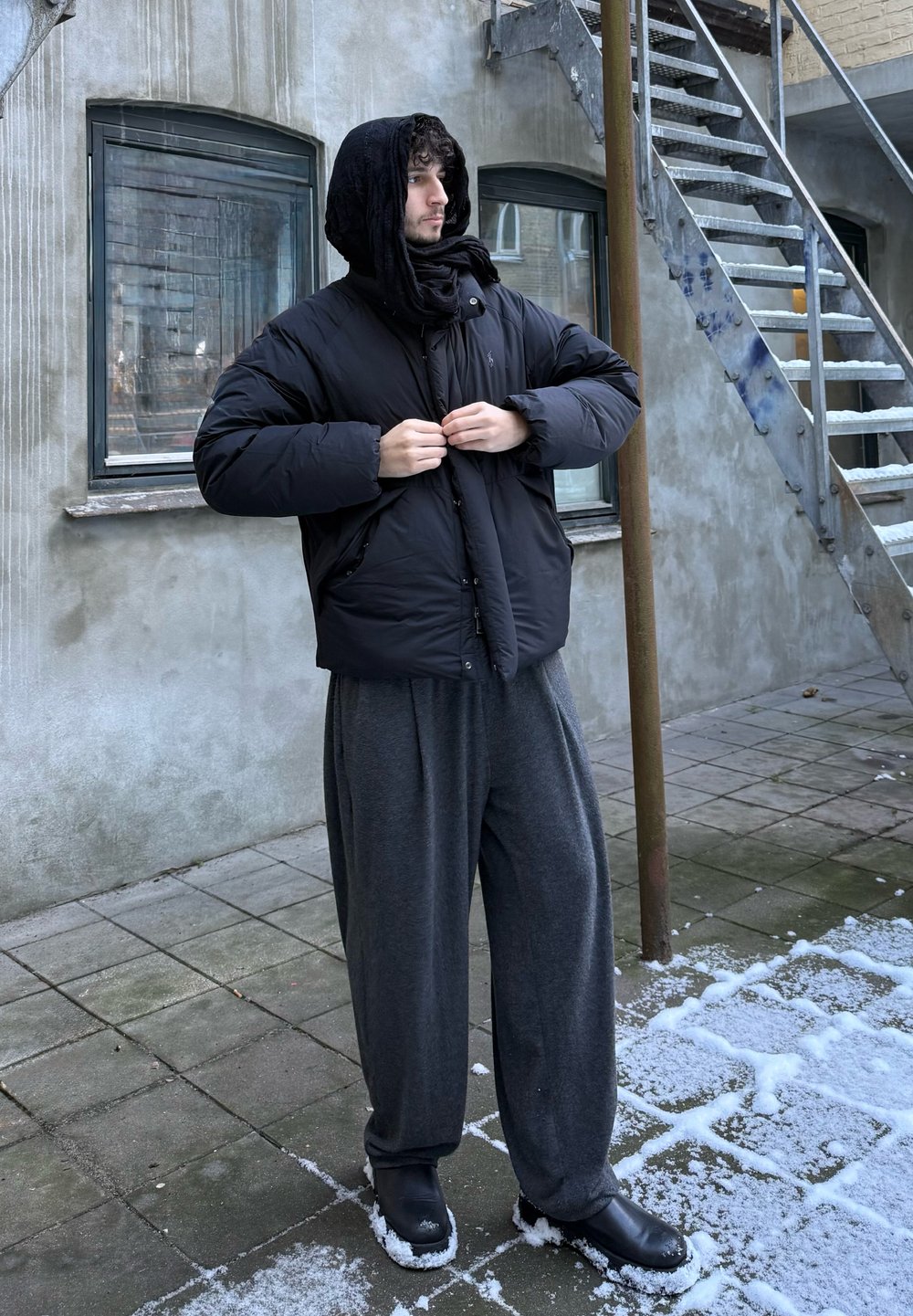 Doudoune noire avec capuche, associée à un pantalon gris ample et des chaussures noires. Le cadre présente un sol enneigé et un mur texturé en arrière-plan.