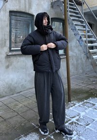 Doudoune noire avec capuche, associée à un pantalon gris ample et des chaussures noires. Le cadre présente un sol enneigé et un mur texturé en arrière-plan.