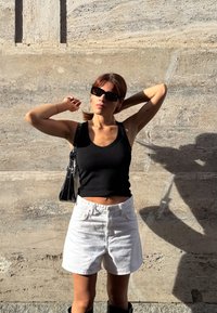 Kvinne i svart tanktop, hvite shorts, solbriller og skulderveske justerer håret mot en solfylt steinvegg.