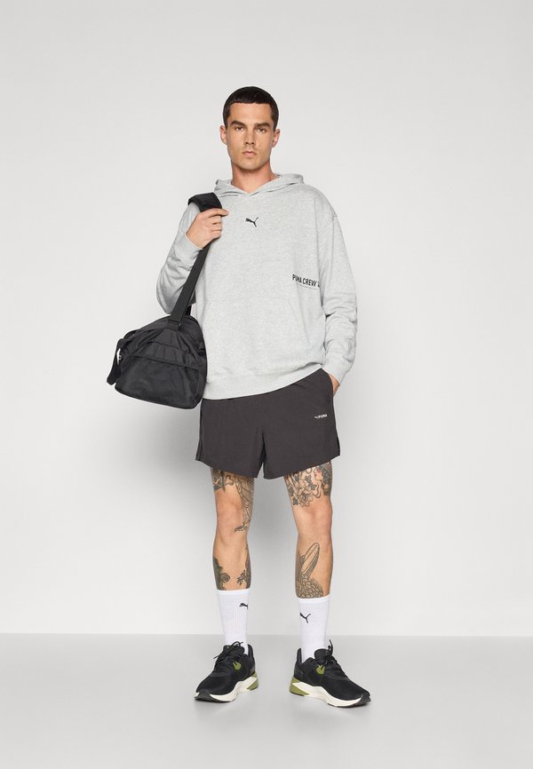 Sudadera gris con el texto "PIMA CREW", pantalones cortos negros con logo, bolsa de deporte negra, zapatillas negras y verdes, calcetines blancos y tatuajes visibles.