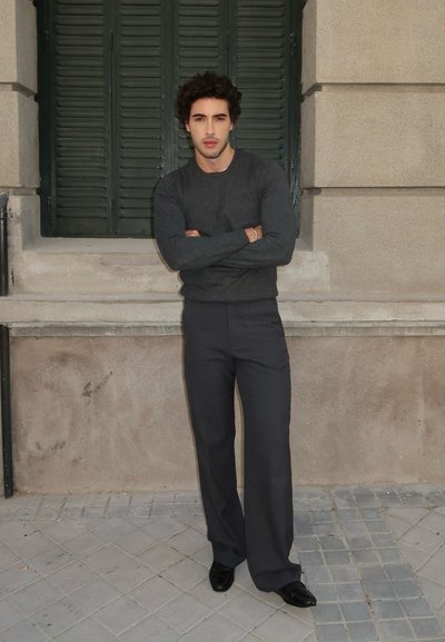 Suéter gris de manga larga, pantalones oscuros ajustados y zapatos negros. De pie contra una pared texturizada con contras verdes al fondo.