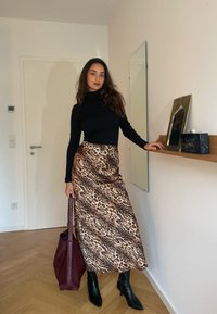 Schwarzer Rollkragenpullover, Midirock mit Leopardenmuster und silbernen Kreiselementen, schwarze Stiefeletten mit Absatz und eine bordeauxfarbene Tote-Bag. Holzboden, minimalistische Dekoration.