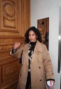 Beige semsket trench coat med store slag, svart genser med et brunt mønster under, holder en kopp, med bakgrunn av trepanel.