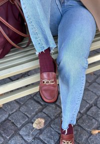 Καφέ δερμάτινες loafers με χρυσές αλ υσίδες συνδυάζονται με βουργουνδί ριγέ κάλτσες και ανοιχτό μπλε τζιν με φθαρμένο στρίφωμα, καθισμένοι σε έναν πάγκο.