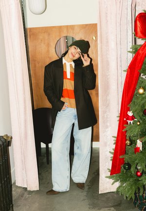 Persona con abrigo negro, suéter a rayas, vaqueros anchos y gorro de lana, de pie entre cortinas rosas junto a un árbol de Navidad decorado.