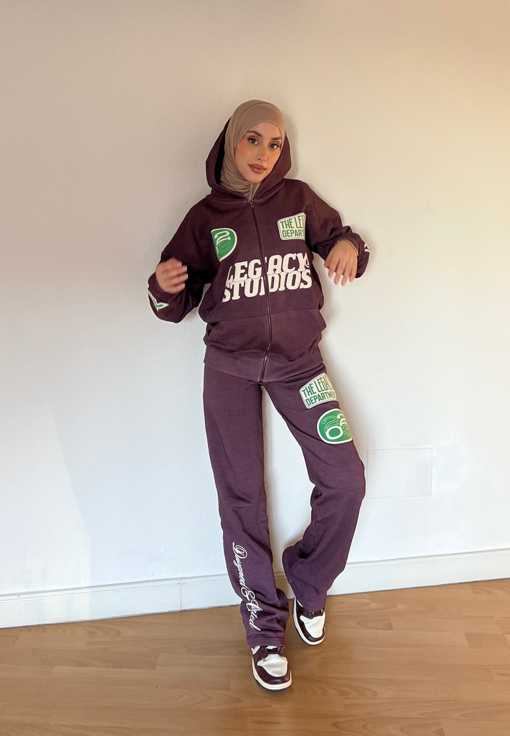 Conjunto de fato de treino roxo que inclui um hoodie com fecho e umas calças a condizer, apresentando texto em branco em destaque e detalhes em verde no logótipo. Usado com ténis brancos e castanhos.