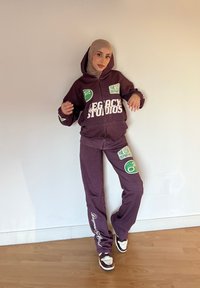 Conjunto de fato de treino roxo que inclui um hoodie com fecho e umas calças a condizer, apresentando texto em branco em destaque e detalhes em verde no logótipo. Usado com ténis brancos e castanhos.