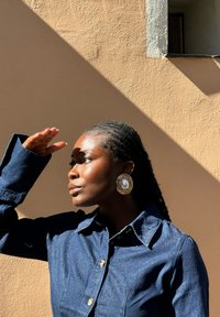 Camicia di jeans con bottoni e colletto, dotata di bottoni dorati. Orecchini grandi e rotondi con un centro simile a una perla e accenti in oro testurizzato. Luce naturale che proietta ombre.
