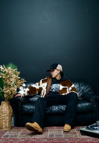 Hombre con chaqueta de piel marrón y blanca, pantalones negros y gorra, sentado en un sofá de cuero negro junto a un jarrón tejido con plantas verdes y beige.