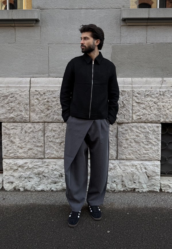 Homme aux cheveux foncés et à la barbe, portant une veste noire zippée et un pantalon large gris à style drapé unique, debout contre un mur en pierre.