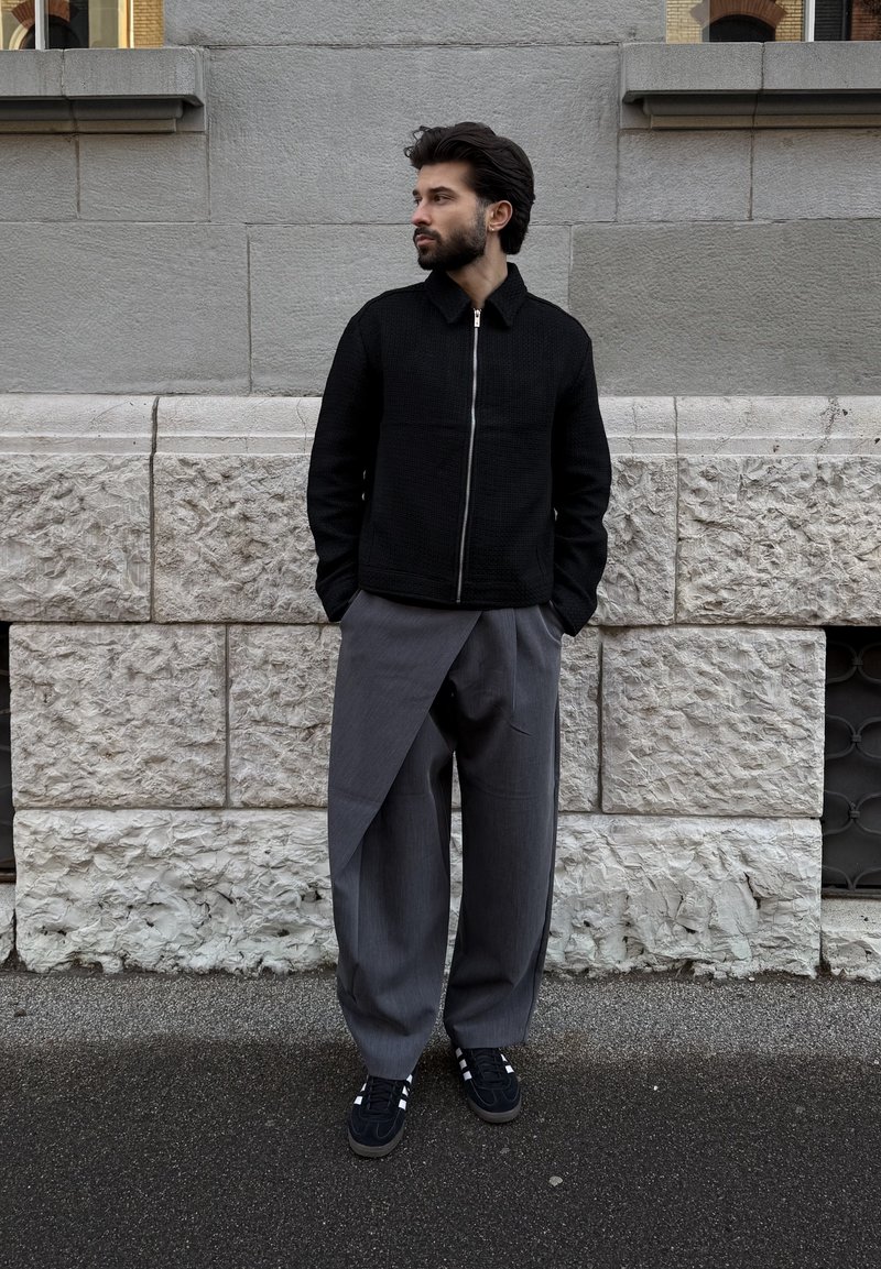 Homme aux cheveux foncés et barbe portant une veste noire zippée et un pantalon large gris unique à style enroulé, debout contre un mur de pierre.