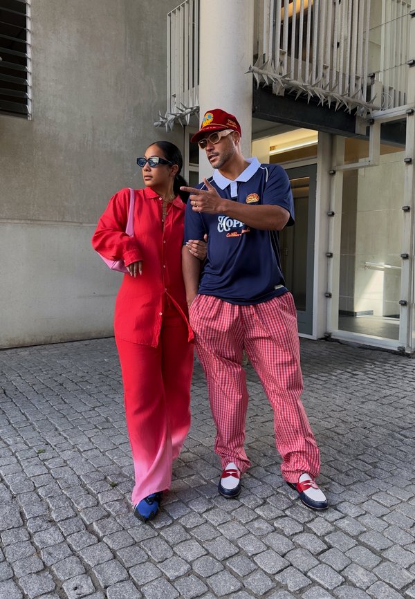 Duas pessoas vestidas à moda atual estão juntas. Uma usa uma camisa de botão vermelha brilhante e calças, enquanto a outra veste uma polo azul-marinho e calças xadrez.