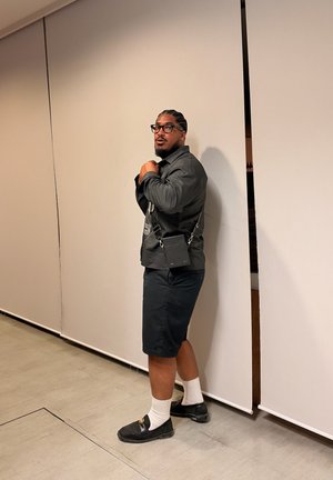 Hombre con el pelo trenzado y gafas, vistiendo chaqueta oscura, pantalones cortos negros, calcetines blancos, zapatos negros y llevando un pequeño bolso negro cruzado dentro de un edificio.
