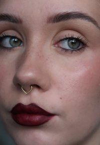 Close-up van een gezicht met een lichte huid, een hartvormige gouden septumring, diepe burgundy matte lippen en goed gedefinieerde wenkbrauwen. Groene ogen met subtiele wimpers.