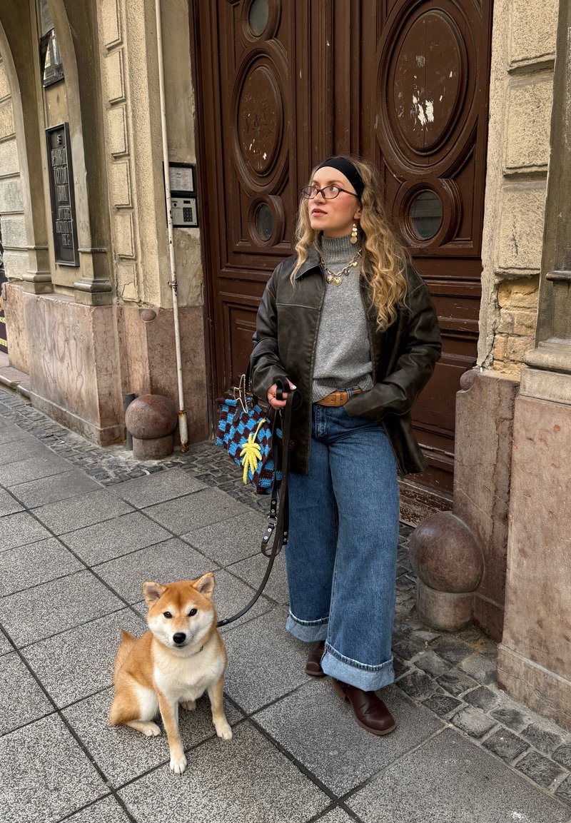 Une personne portant une veste en cuir noire, un col roulé gris et un jean en denim à jambes larges tient une laisse attachée à un Shiba Inu. Le chien est assis sur le trottoir.