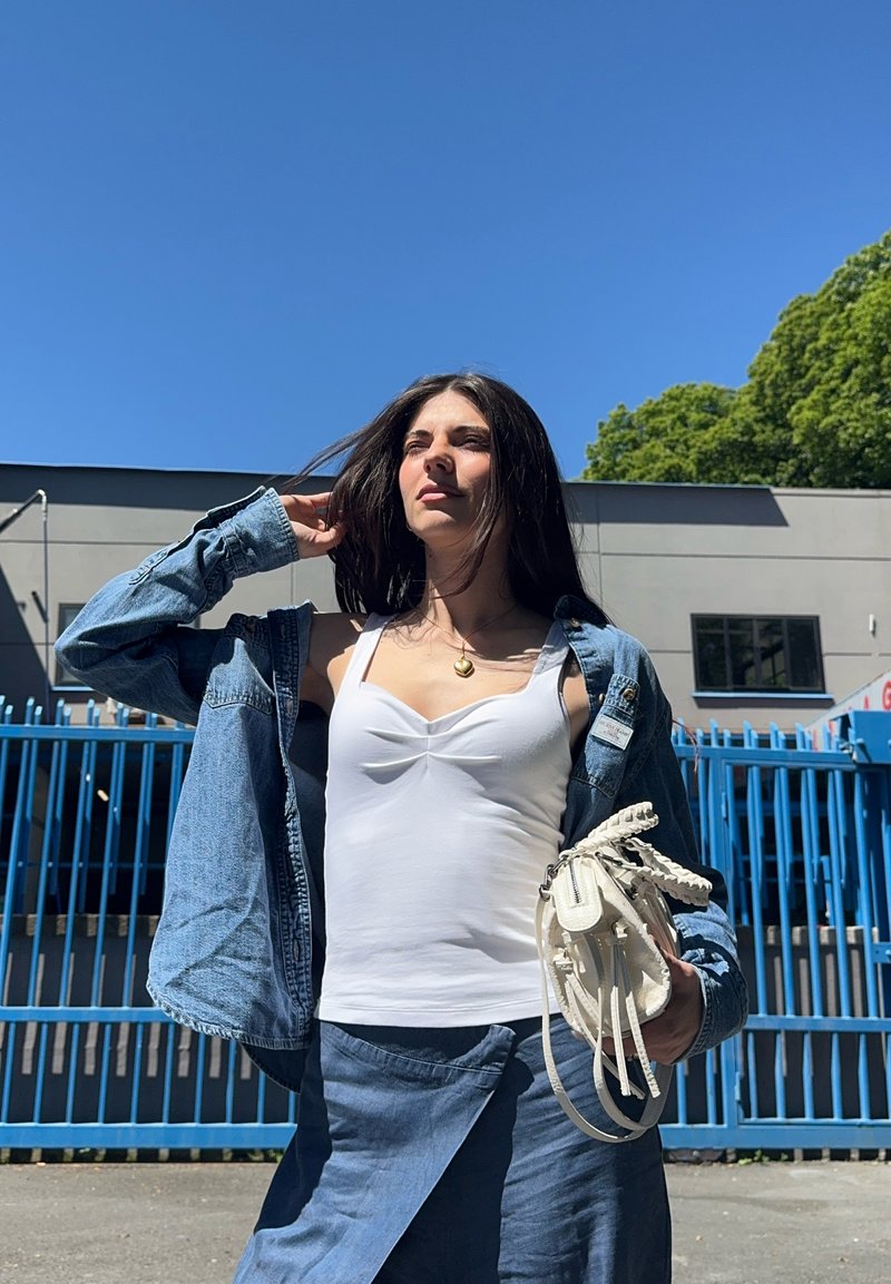 Donna con canotta bianca e giacca di jeans sta all'esterno tenendo una borsa bianca, con una recinzione blu e un edificio dietro sotto un cielo azzurro limpido.