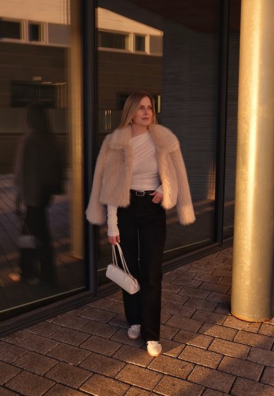 Abrigo de piel sintética en crema, camisa ajustada de manga larga, pantalones negros de tiro alto, zapatillas blancas y un pequeño bolso blanco de hombro.