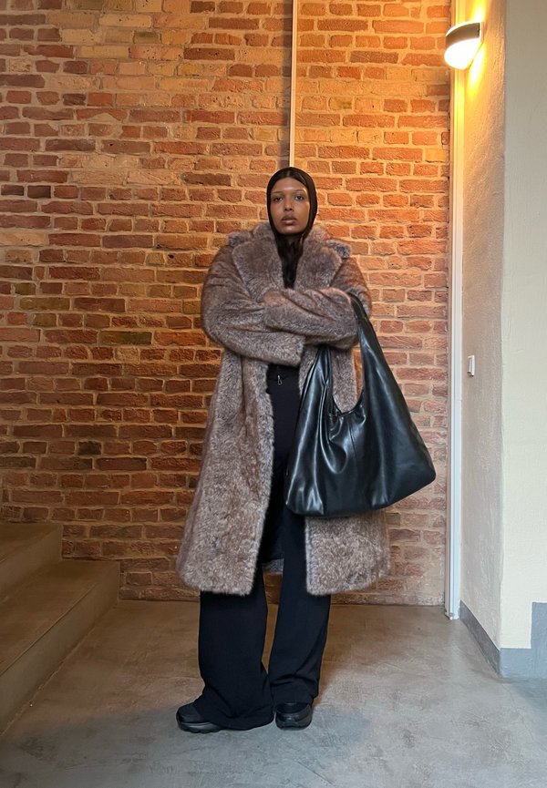 Langer, brauner Kunstfellmantel mit großem Kragen, kombiniert mit schwarzer Kleidung und einer schwarzen Lederhandtasche in einem Innenraum mit Ziegelwänden.