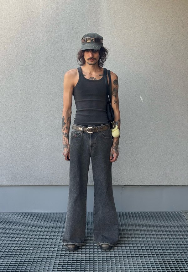 Jeune personne aux longs cheveux, tatouages, débardeur noir, jeans gris larges, casquette avec lunettes de soleil sur le dessus, debout contre un mur gris uni.