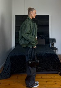 Kobieta w zielonej kurtce i czarnych szerokich spodniach stoi bokiem w sypialni, trzymając małą czarną torebkę, za nią znajduje się łóżko i lampa.