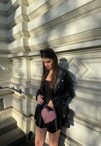 Svart krokodilstrukturerad läderjacka över en svartrandig klänning, hållande en rosa hjärtformad väska med kedjestropp. Solbelyst vägg i bakgrunden.