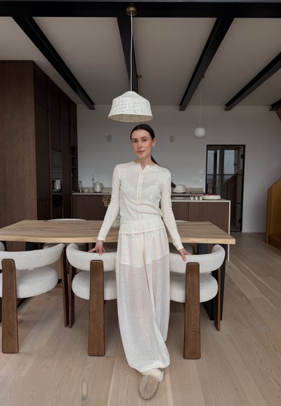 Mujer con blusa blanca de manga larga y pantalones sueltos apoyada en una mesa de comedor de madera con sillas tapizadas en un interior de cocina moderna.