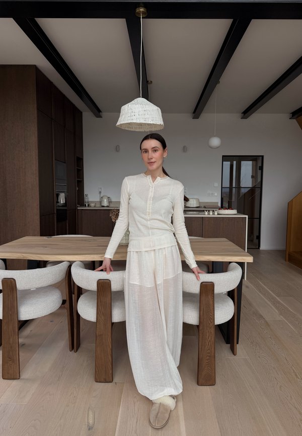 Mujer con blusa blanca de manga larga y pantalones sueltos apoyada en una mesa de comedor de madera con sillas tapizadas en un interior de cocina moderna.