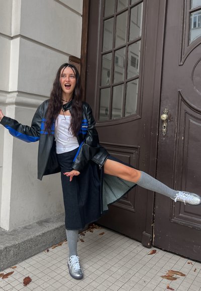 Chaqueta de cuero negra y azul, blusa blanca, falda gris, calcetas gris hasta la rodilla y zapatillas plateadas. Persona adoptando una pose juguetona cerca de una puerta.
