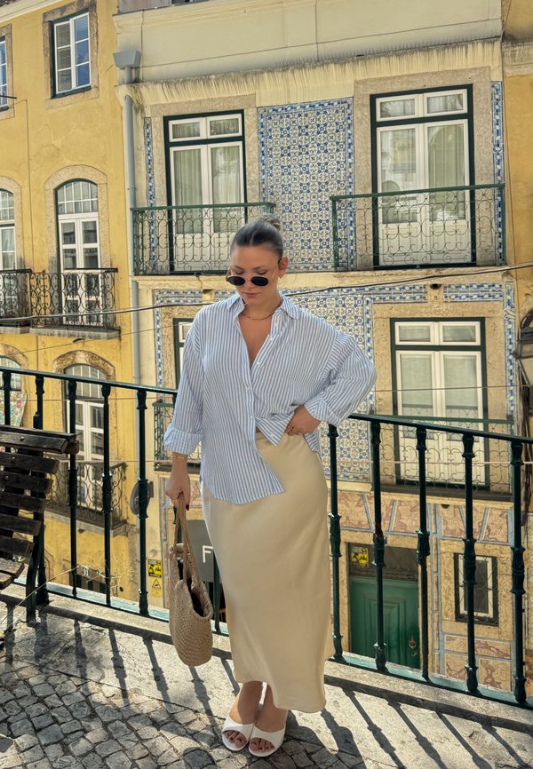 Femme portant des lunettes de soleil, une chemise rayée, une jupe beige et des sandales blanches, debout sur un balcon avec en arrière-plan des bâtiments décorés de carreaux.