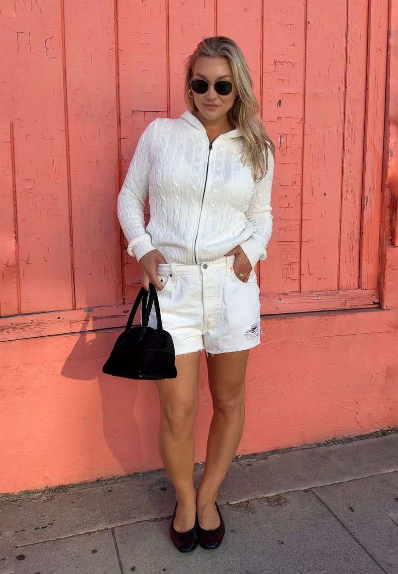 Femme portant un pull zippé blanc et un short blanc, tenant un sac à main noir, debout contre un mur en bois corail sur le trottoir.