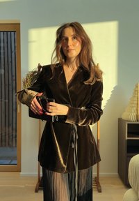 Femme en blazer en velours noir et jupe plissée tenant un petit sac noir, debout sous une lumière chaude dans une pièce moderne.