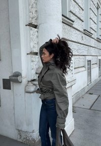 Geacă verde măsliniu cu curea și guler înalt, asortată cu blugi albastru închis. Modelul ține o geantă maro texturată, stând lângă o clădire.