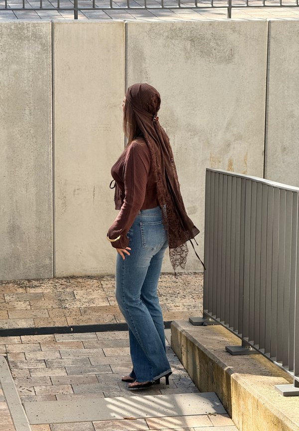 Kvinde iført et brunt tørklæde, langærmet top, blå jeans og hæle, står ved siden af et rækværk på en stensat sti.