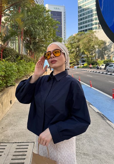 Mujer con un pañuelo blanco en la cabeza, chaqueta negra oversize y gafas de sol con tinte amarillo, de pie en la acera de la ciudad sosteniendo una bolsa de papel.