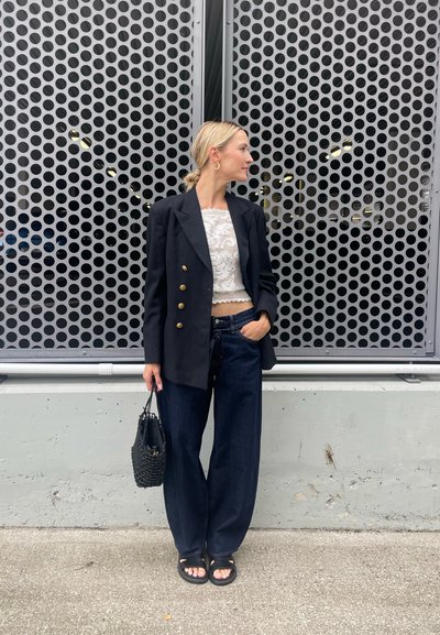 Blazer negro de doble botonadura sobre un top corto de encaje blanco, combinado con jeans anchos oscuros y un bolso negro de mimbre. El fondo presenta metal perforado.