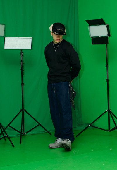 Camisa negra de manga larga, pantalones vaqueros azul oscuro, zapatos grises; lleva un sombrero negro y beige; luces de estudio en trípodes; fondo verde.