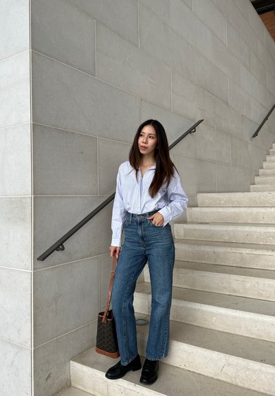 Camiseta blanca a rayas de manga larga, vaqueros acampanados de mezclilla azul de cintura alta, zapatos de plataforma negros, llevando un bolso tipo bucket negro y marrón, de pie en las escaleras.