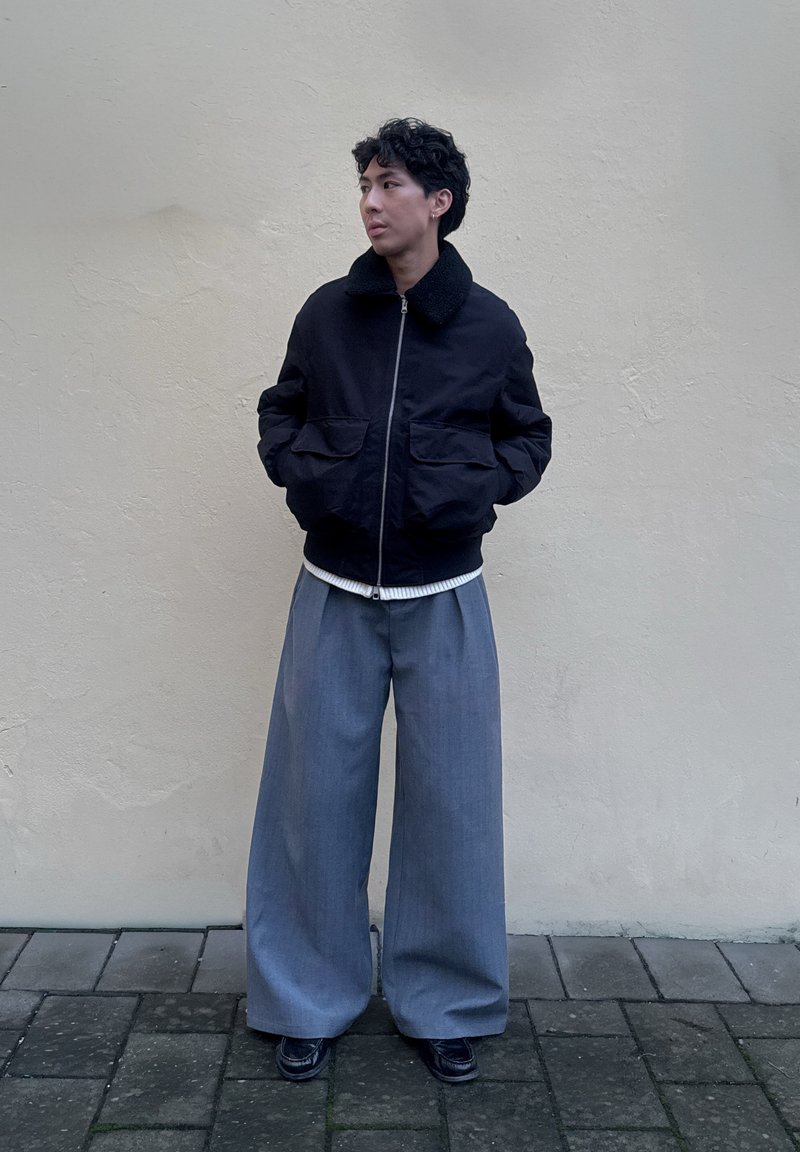 Schwarze Bomberjacke mit Pelzkragen, zwei Fronttaschen; graue weite Hosen; schwarze Schuhe; vor einer neutralen Wand stehend.