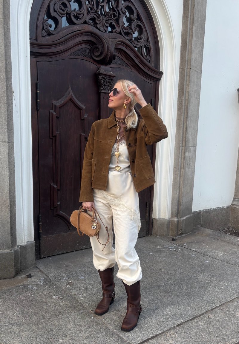 Femme en veste en daim marron, pantalon blanc et bottes foncées, tenant un sac à main beige devant une porte en bois foncé ornée.