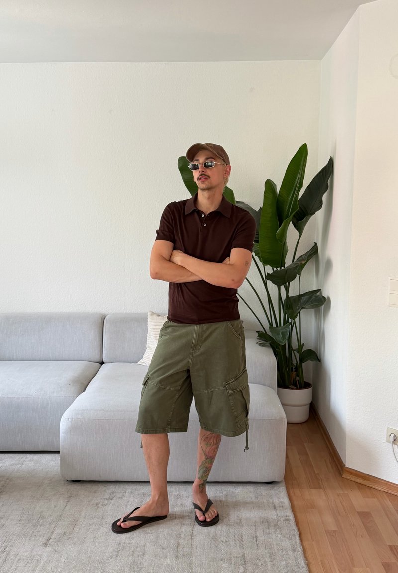 Homme portant des lunettes de soleil, une chemise marron, un short vert et des tongs, debout les bras croisés dans un salon à côté d’un canapé gris et une grande plante.