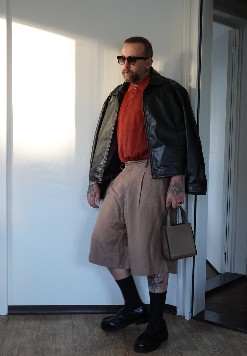 Mand med tatoveringer bærer sort læderjakke over rustfarvet skjorte og beige vide shorts, holder lille beige håndtaske indendørs nær døren.