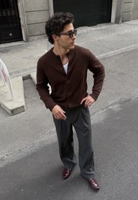 Donkerbruine knoop-up cardigan, witte t-shirt, grijze broek met krijtstrepen en bordeauxrode schoenen. Glad textuur met een relaxte pasvorm.