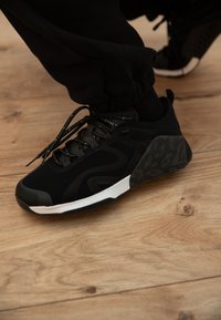 Personne portant des baskets à motifs noirs et gris avec des semelles intermédiaires blanches, debout sur un sol en bois clair.