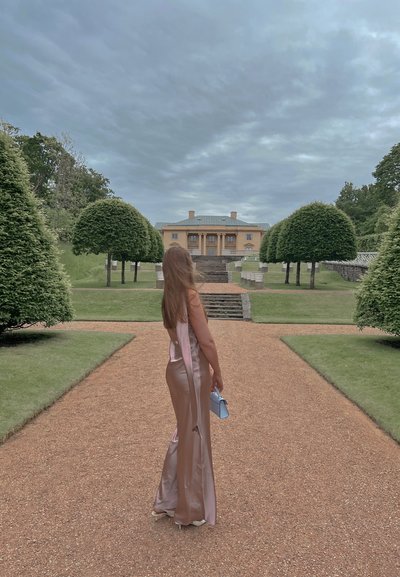 Largo vestido de satén color rubor con una abertura alta; modelo sosteniendo un bolso azul claro, de pie en un camino de grava hacia un edificio neoclásico.